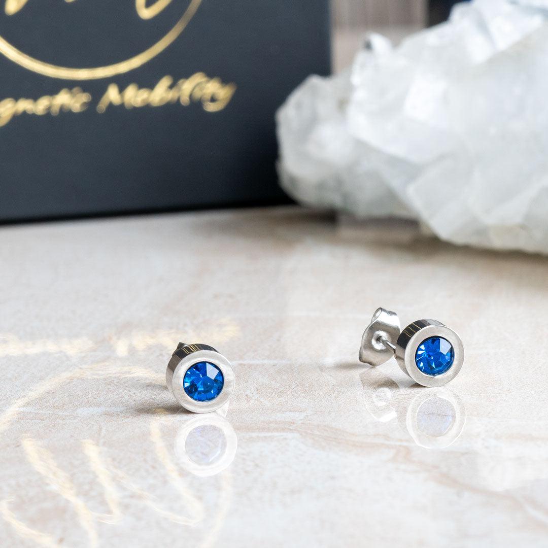 Birthstone Stud Earrings by Magnetic Mobility with blue Swarovski crystals. These earrings have a magnetic back and are ideal for fashionable pain relief, displayed on a reflective surface with Magnetic Mobility packaging.