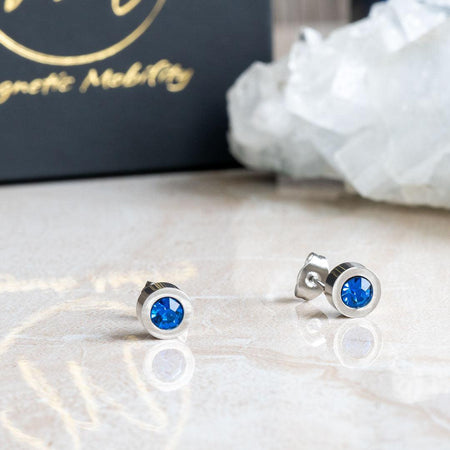 Birthstone Stud Earrings by Magnetic Mobility with blue Swarovski crystals. These earrings have a magnetic back and are ideal for fashionable pain relief, displayed on a reflective surface with Magnetic Mobility packaging.